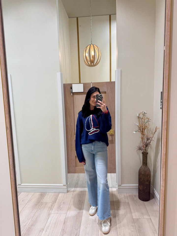 Person taking a mirror selfie wearing a blue sweater with red and white details, light blue jeans, and white sneakers.