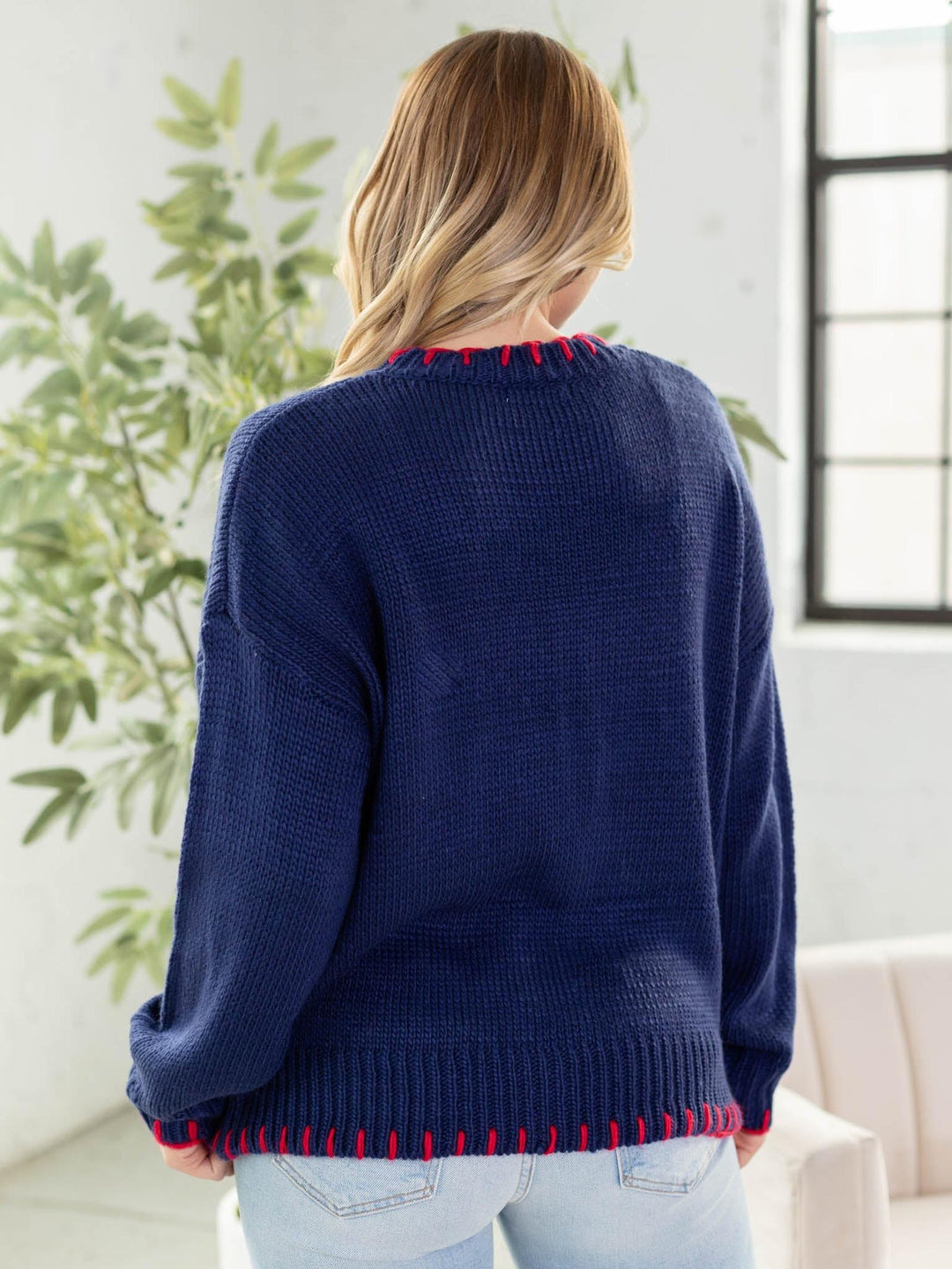 Person wearing a navy sweater with red trim in a room with plants and a window.