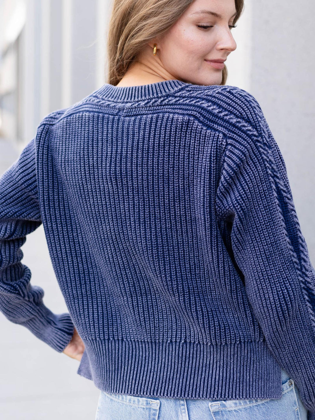 A woman wearing a navy blue ribbed knit sweater, shown from the back and side. The sweater features a textured cable-knit pattern around the neckline and shoulders, ribbed cuffs and hem, and is paired with light blue jeans.