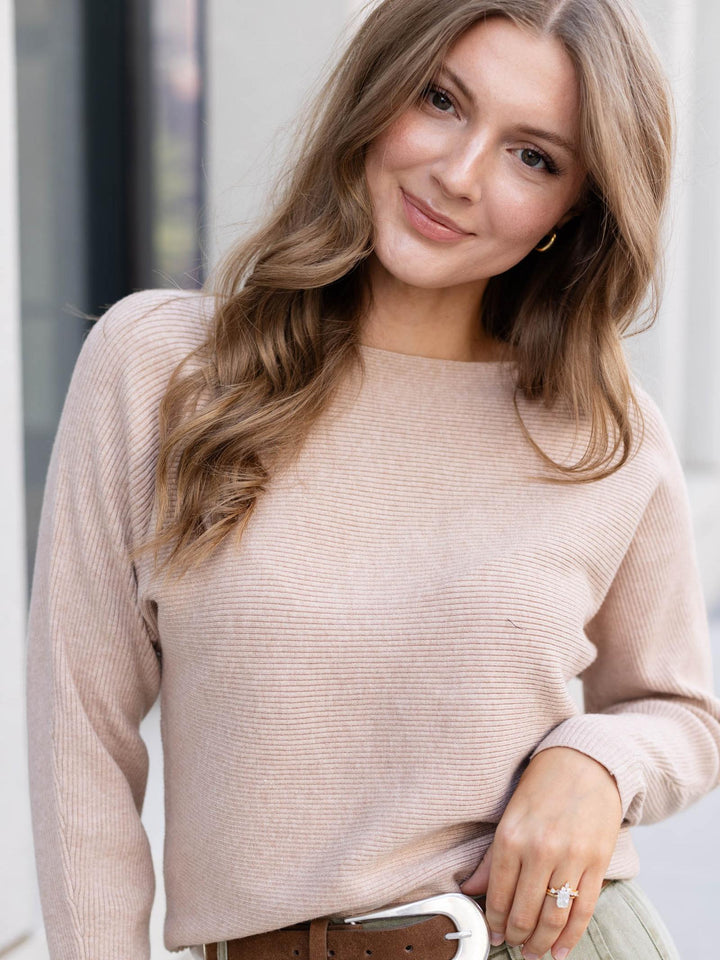 A woman wearing a light beige ribbed knit sweater, styled with a brown belt and light olive pants, photographed in a bright, modern setting.