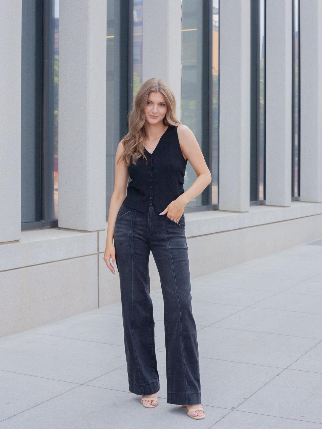 A woman stands outdoors in front of a modern building with large windows and concrete columns, wearing a sleeveless black V-neck top and dark wide-leg jeans.
