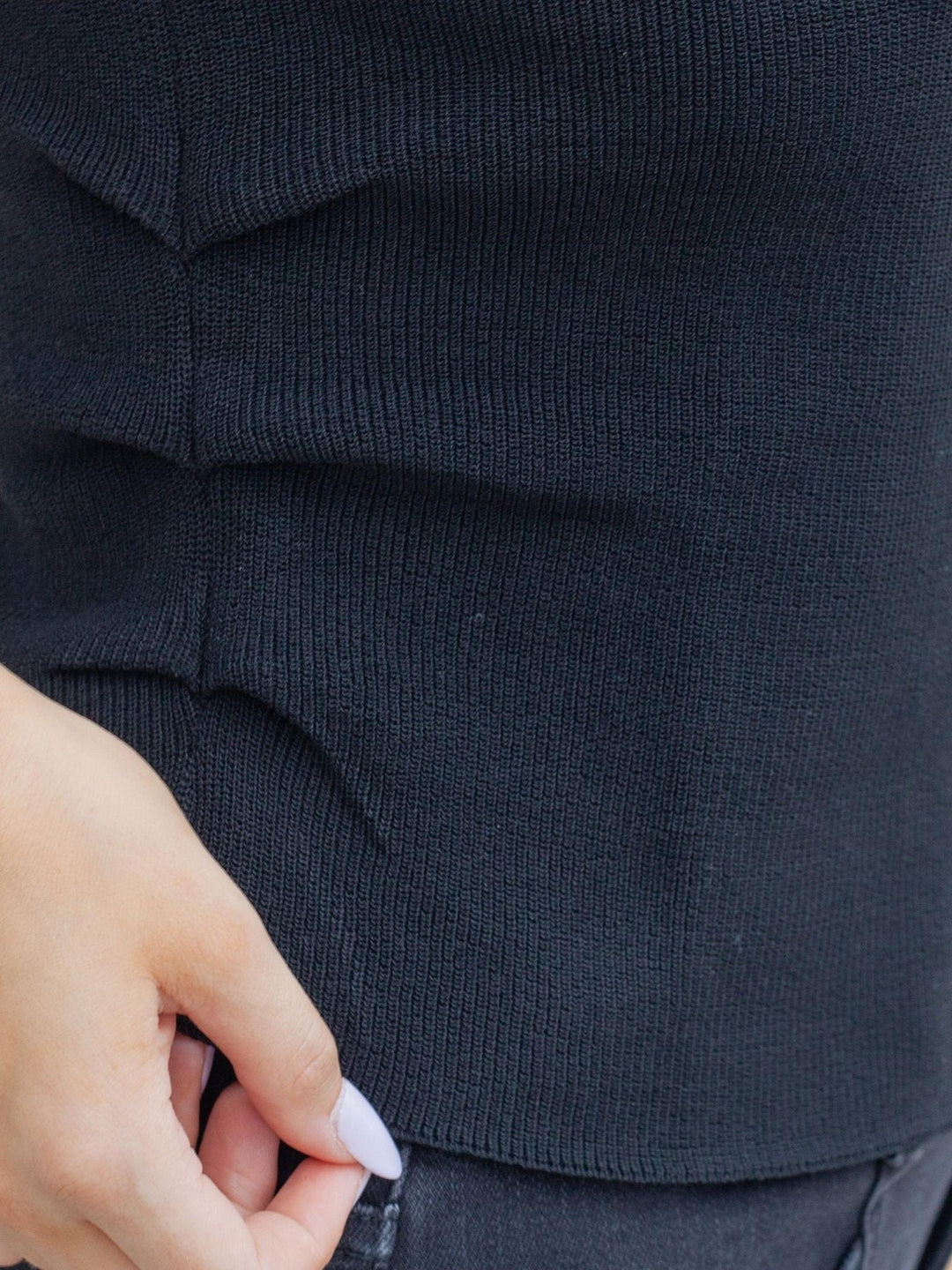 Close-up of a person wearing a black ribbed knit top, with a hand pulling the hem slightly.