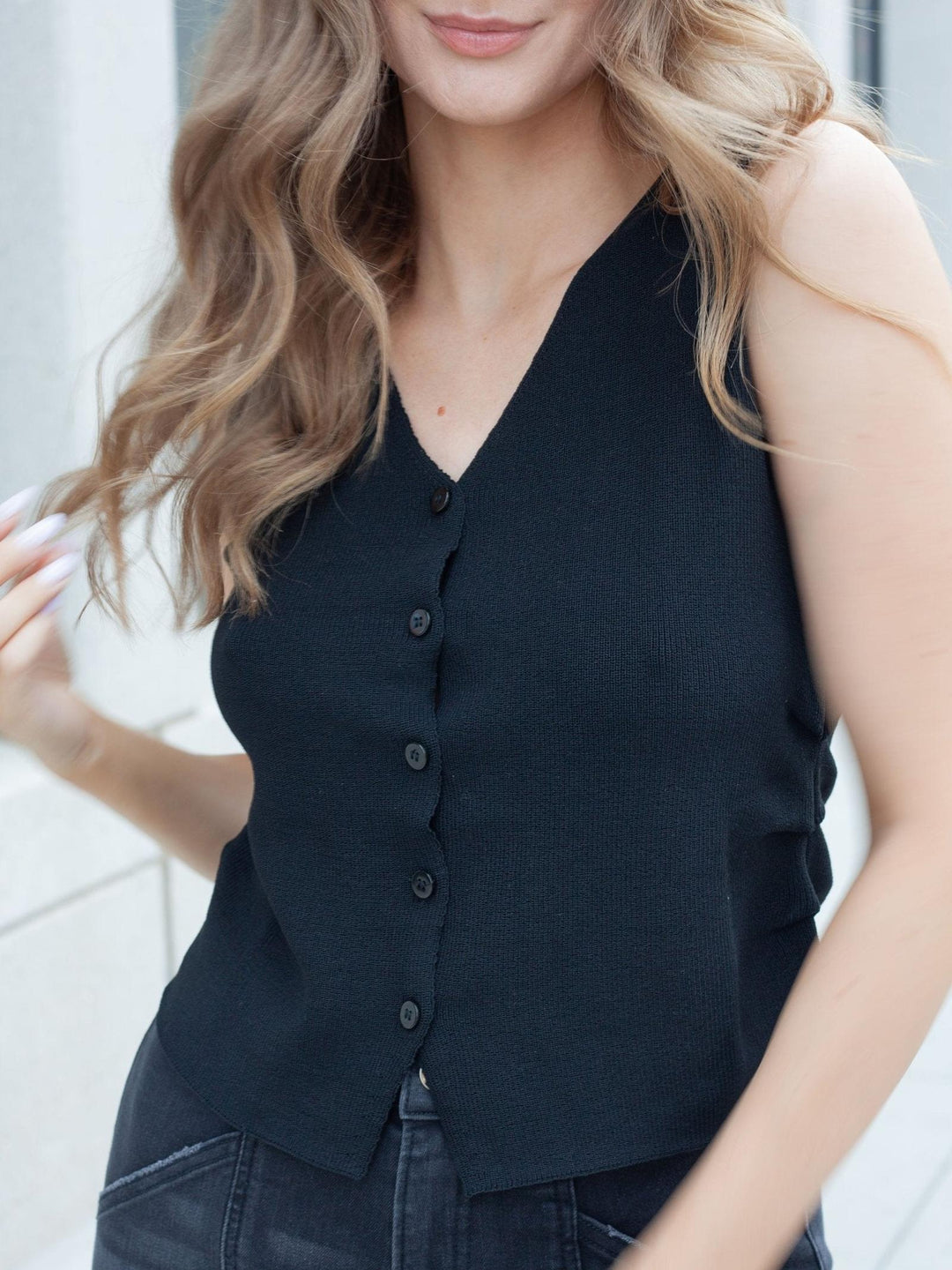 A woman wearing a sleeveless black V-neck top with a button-front closure, paired with dark jeans. The photo is cropped from the lower face to the hips, set against a softly blurred light gray outdoor background.