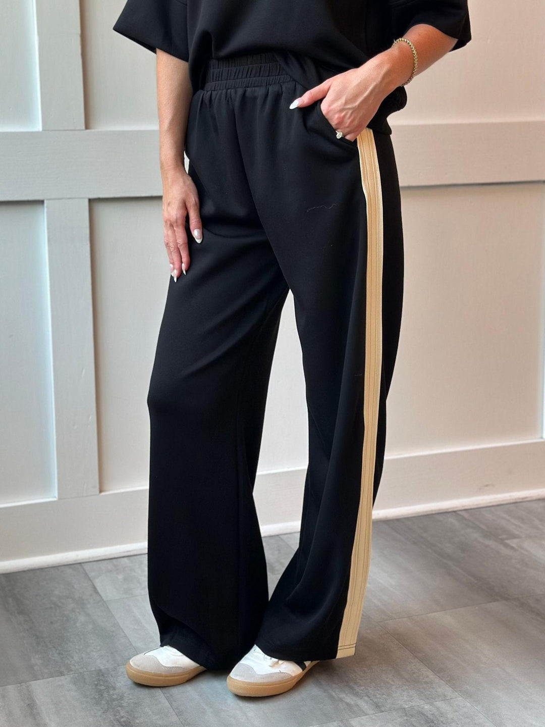 A person wearing black wide-leg pants with a beige side stripe, paired with a black top and white sneakers, standing indoors against a paneled wall.