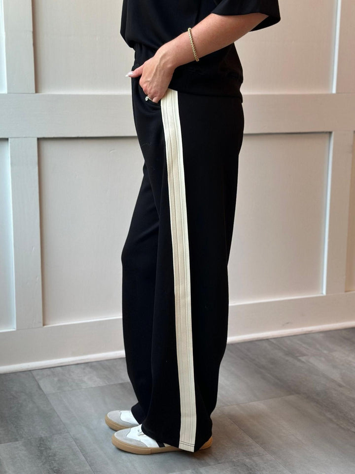 A person wearing black wide-leg pants with a prominent off-white pleated vertical stripe down the side, paired with a black top and white sneakers, standing in front of a white paneled wall.