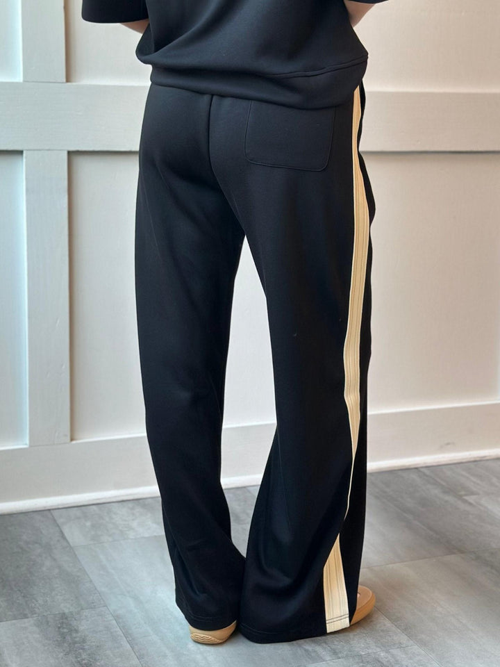 Black wide-leg sweatpants with a vertical beige stripe down the side, shown on a model from the back in an indoor setting.