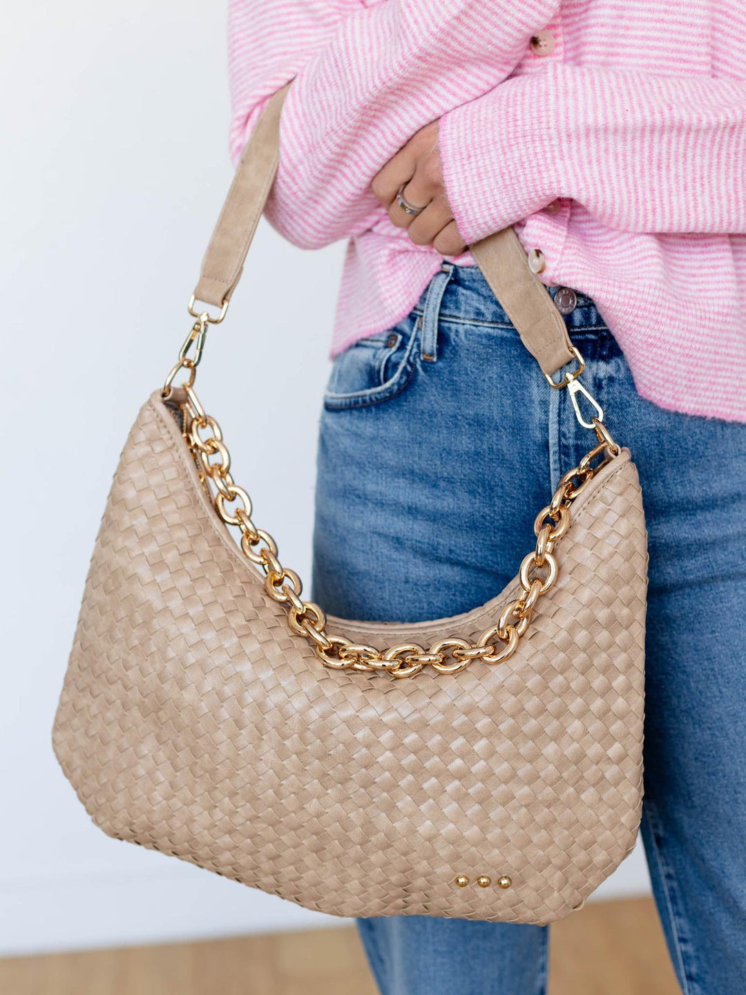 Person holding a beige woven handbag with a gold chain detail, wearing a pink sweater and blue jeans.