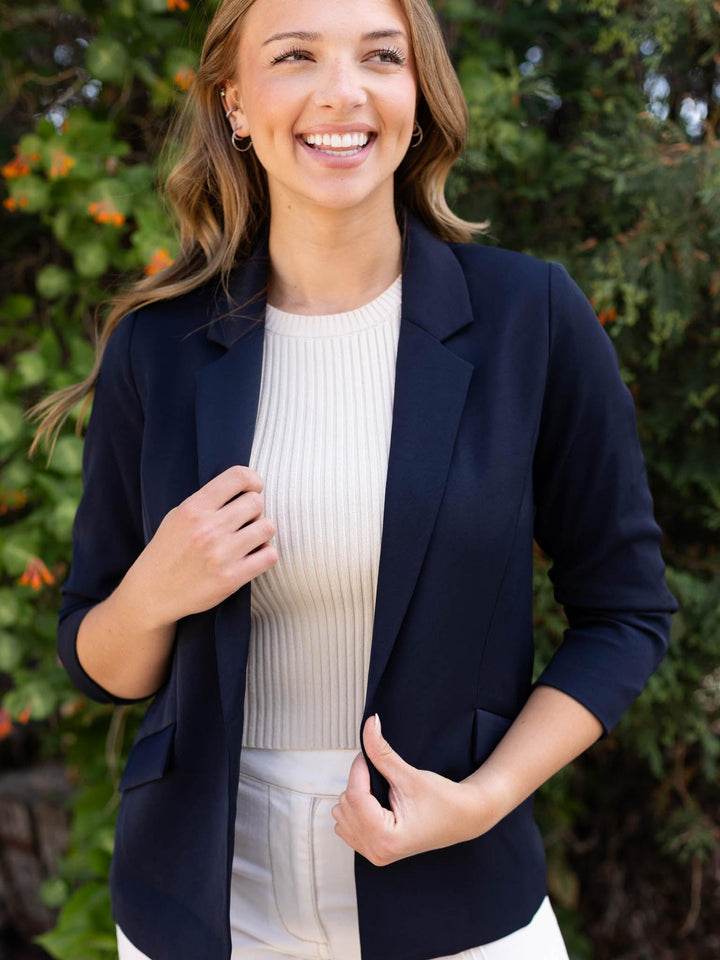 Smiling woman in a navy recycled shirred sleeve blazer over a cream ribbed top, outdoors.