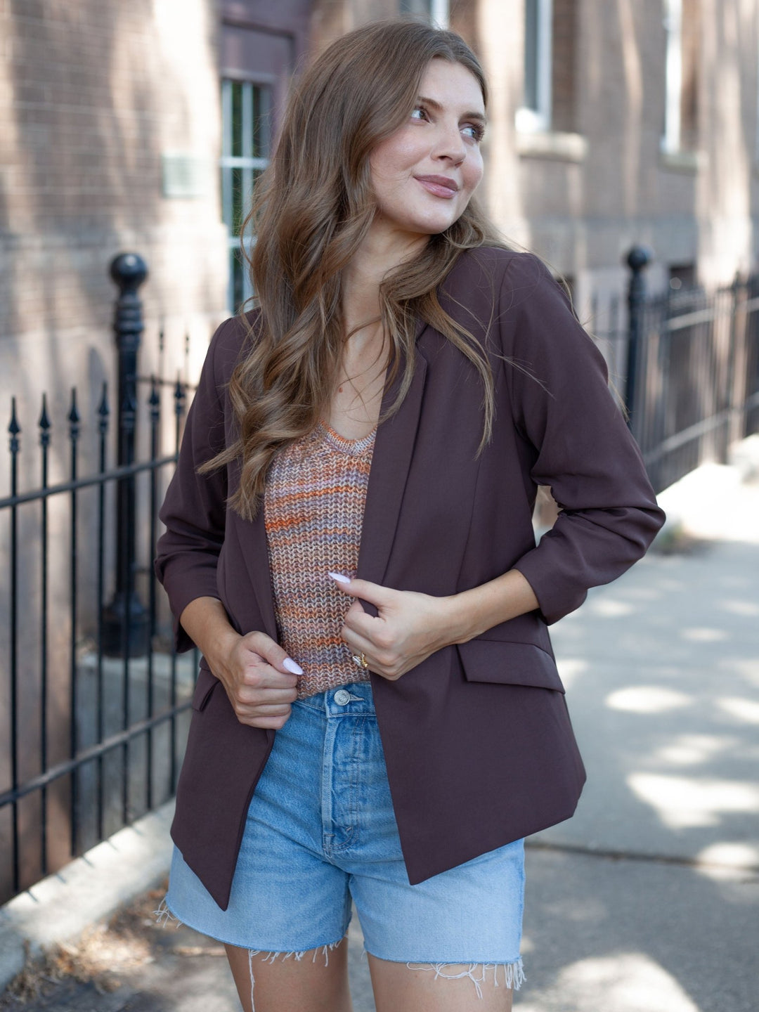 A woman wearing a dark plum purple blazer over a patterned orange and beige top, paired with light blue denim shorts, standing on a city sidewalk.