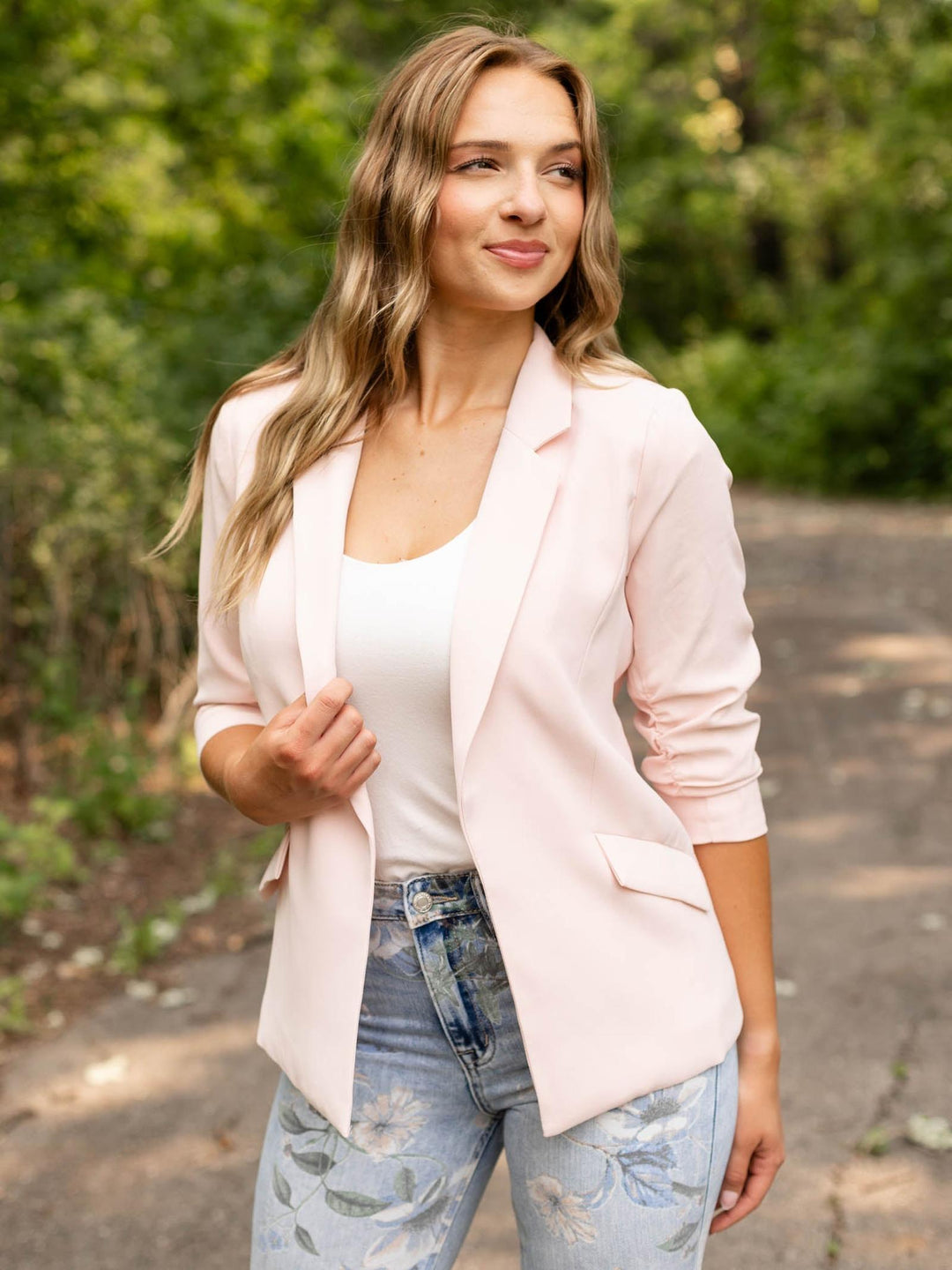 A woman wearing a light pink blazer over a white tank top and light blue floral jeans, standing outdoors on a paved path with green foliage in the background.