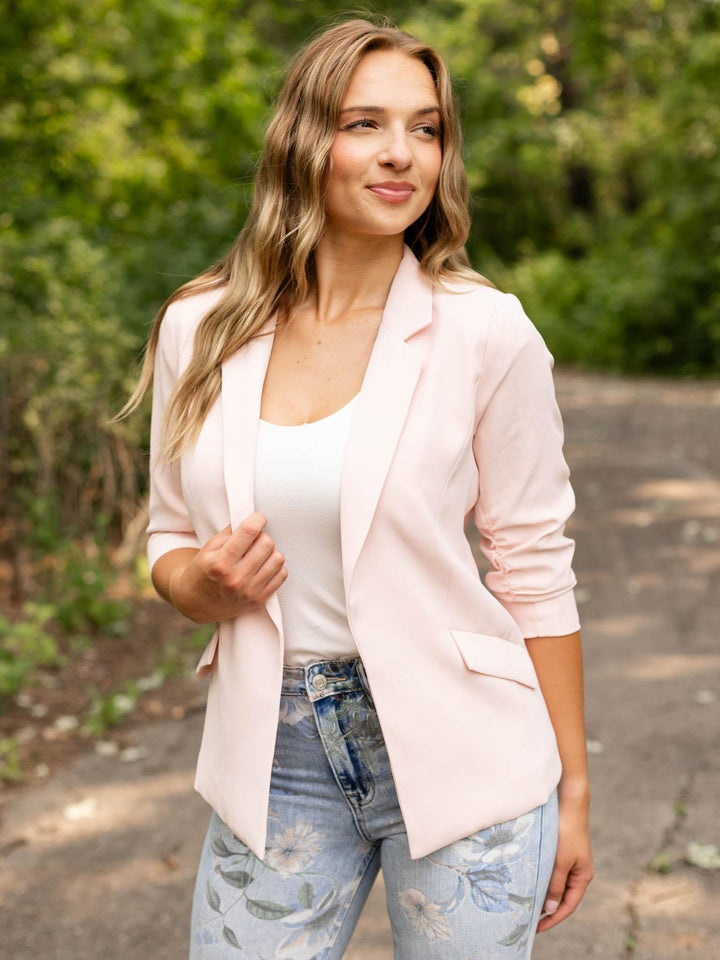 A woman wearing a light pink blazer over a white tank top and light blue floral jeans, standing outdoors on a paved path with green foliage in the background.