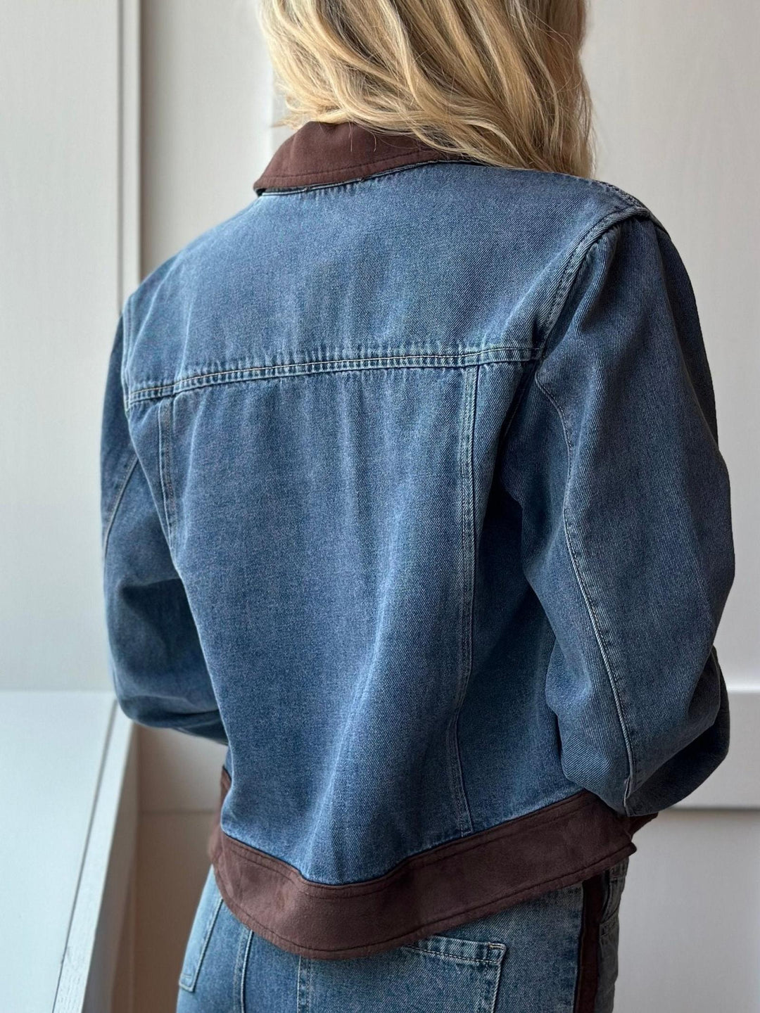A person shown from the back wearing a cropped blue denim jacket with contrasting dark brown suede-like trim at the collar and hem, paired with blue jeans in a bright indoor setting.
