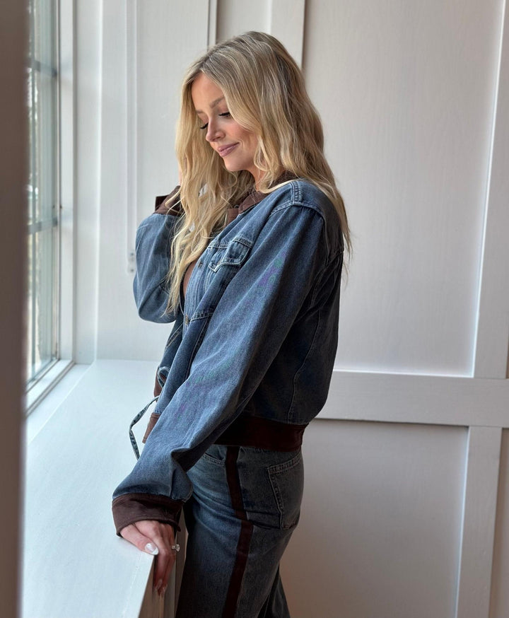 A woman with blonde hair stands by a window wearing a blue denim jacket with brown contrast trim and matching blue jeans with brown side stripes.