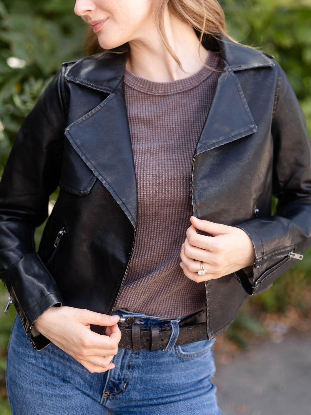 A woman wearing a black faux-leather moto jacket over a brown ribbed knit sweater and blue jeans, photographed outdoors with a blurred green background.