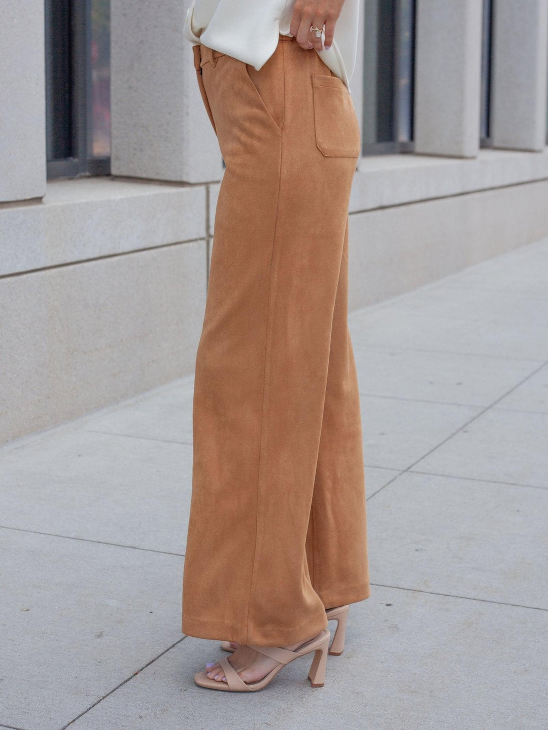 A person wearing wide-leg tan suede pants, a white top, and beige heeled sandals, standing on a light concrete sidewalk in front of a modern building.