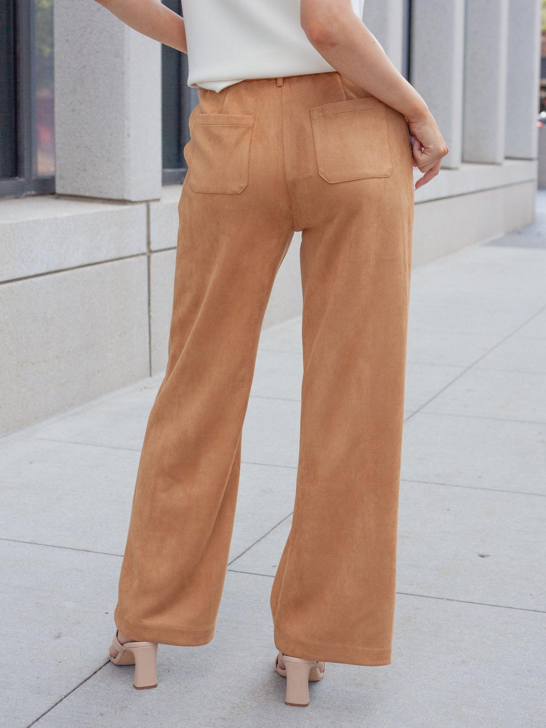 A person shown from the back wearing tan wide-leg pants with a white top and beige heeled sandals, standing on a light concrete sidewalk.