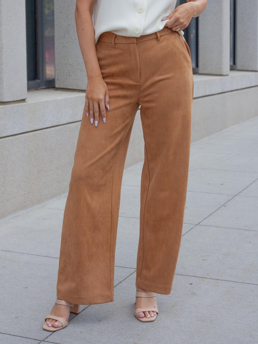 A person wearing tan wide-leg trousers with a white blouse and nude heeled sandals, standing on a concrete sidewalk in front of a modern building.