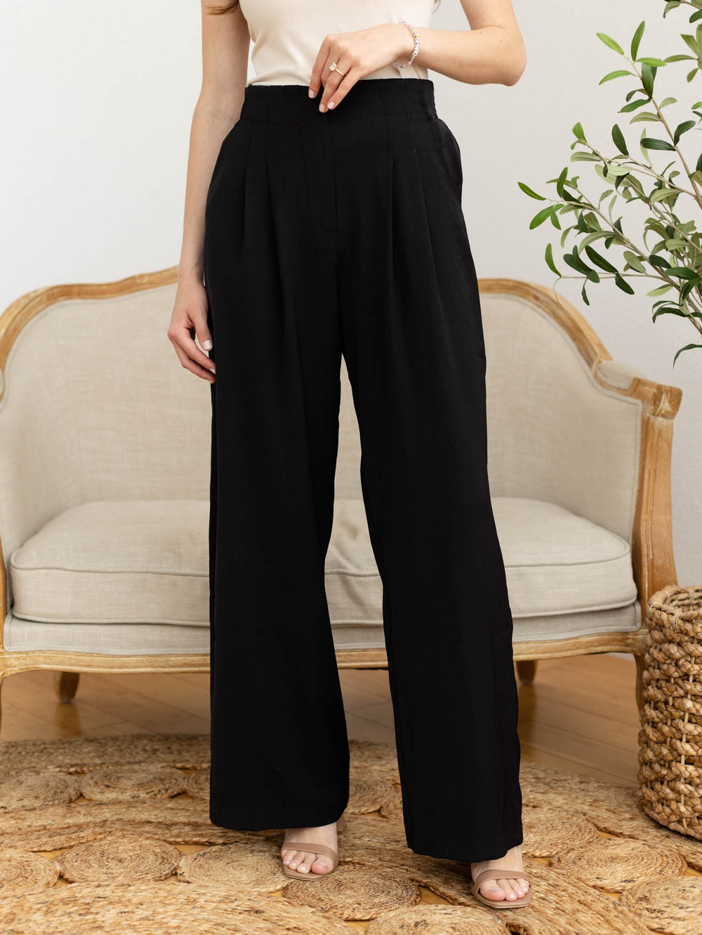 Person wearing black wide-leg pants standing in a room with a beige armchair and plant.