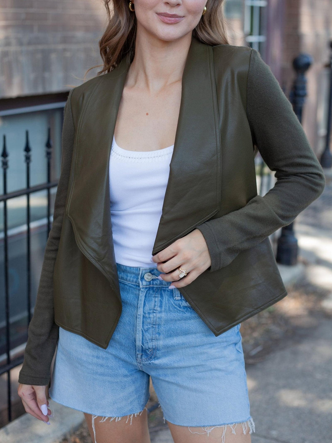 A woman wearing an olive green faux-leather blazer over a white tank top and light-wash denim shorts, standing outdoors on a sidewalk.