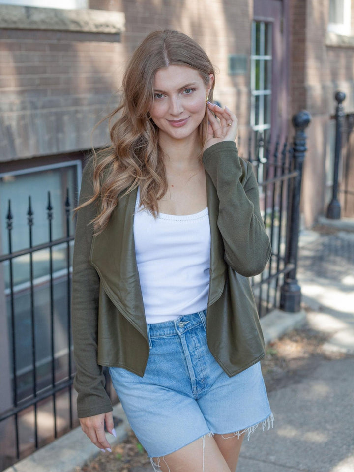 A woman wearing an olive green wrap-style jacket over a white tank top and light blue denim shorts, standing outdoors on a sidewalk in front of a brick building and black metal fence.
