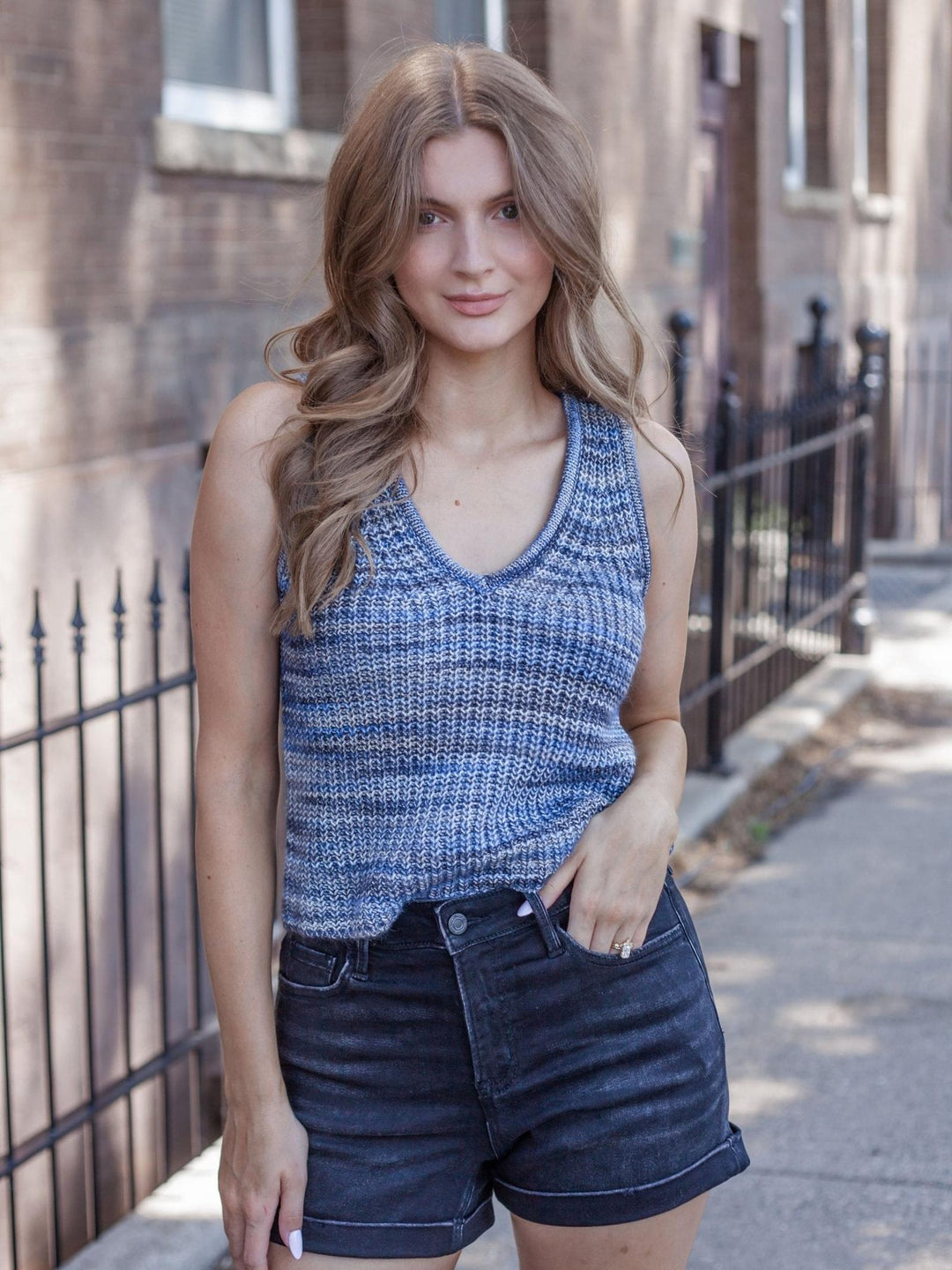 A woman standing outdoors on a sidewalk wearing a blue and gray textured knit sleeveless tank top and dark denim shorts.