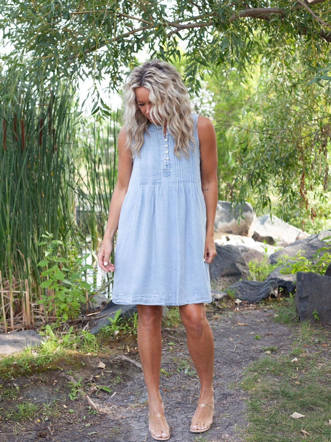A woman wearing a light blue sleeveless button-front dress stands outdoors in a garden setting with trees, plants, and rocks in the background.