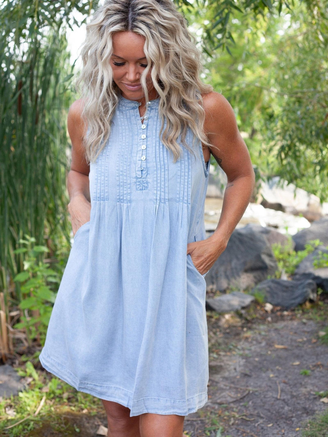 A woman wearing a light blue sleeveless denim dress with a buttoned neckline and side pockets, standing outdoors in a garden setting.