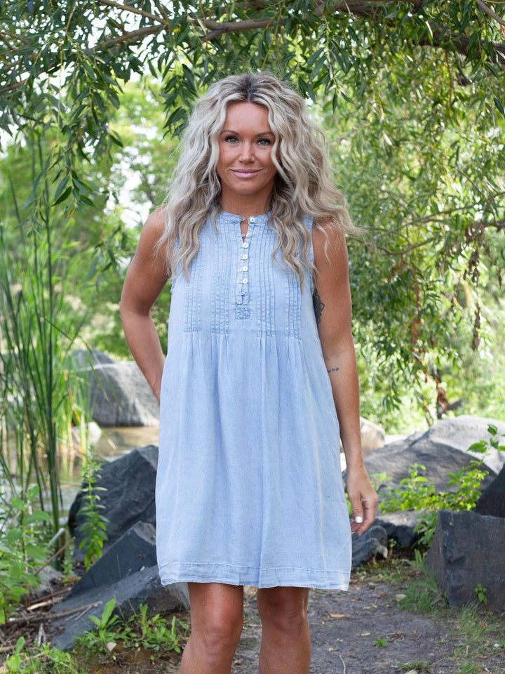 A woman with long blonde hair wearing a light blue sleeveless linen dress, standing outdoors in a garden setting with rocks and green foliage.
