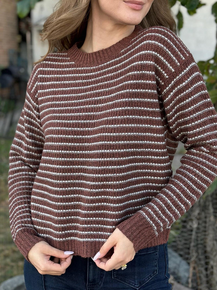 A woman wearing a brown and white horizontally striped knit sweater, paired with dark blue jeans, photographed outdoors with a blurred natural background.