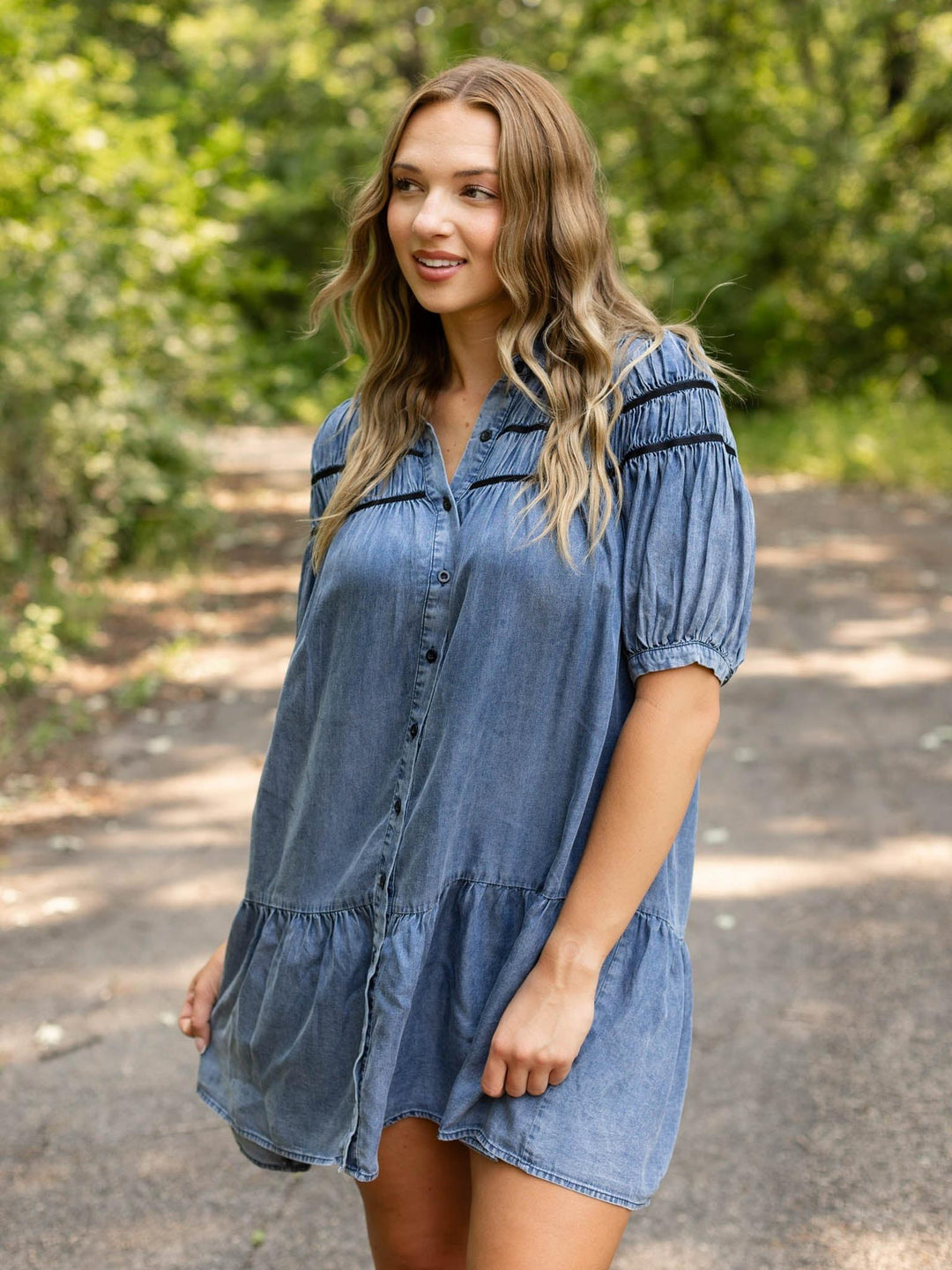 A woman wearing a blue denim short-sleeve button-up dress with puff sleeves and a ruffled hem, standing outdoors on a path with trees in the background.