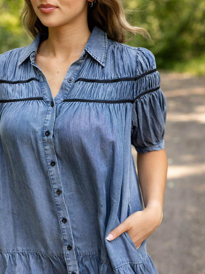 A woman wearing a blue denim-like short-sleeve button-up shirt with black horizontal trim details across the chest and shoulders, photographed outdoors with a blurred natural background.