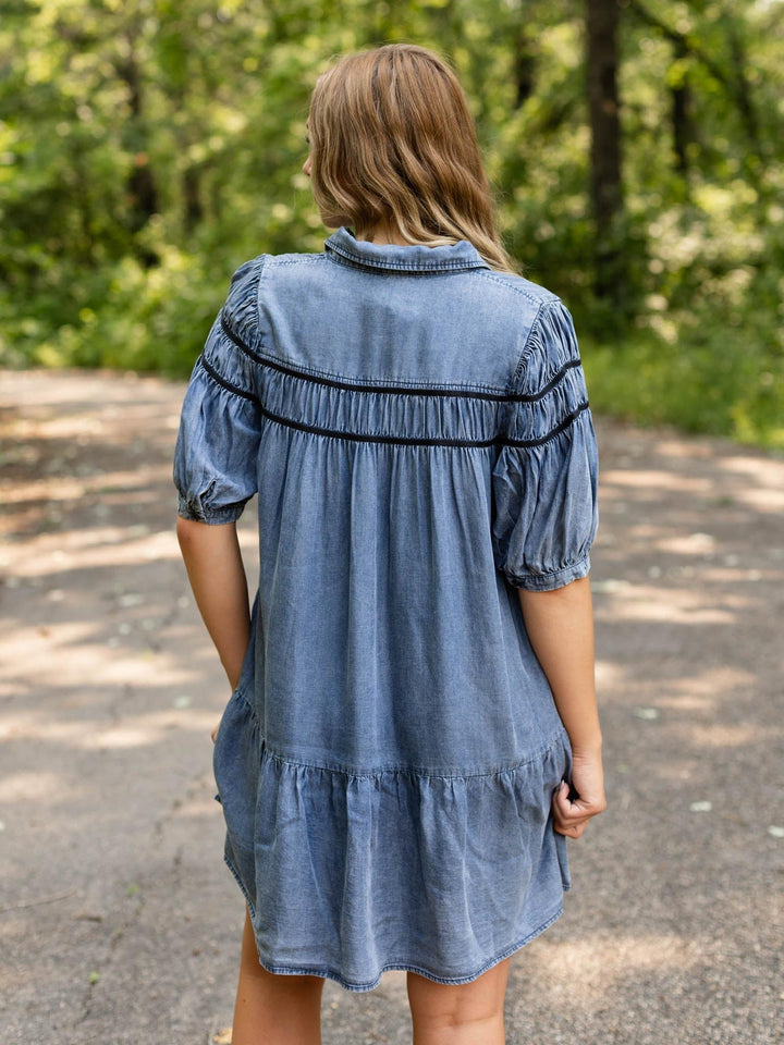 A woman viewed from behind wearing a blue denim short-sleeve dress with black embroidered horizontal stripes across the shoulders, standing on an outdoor paved path with a blurred green forest background.