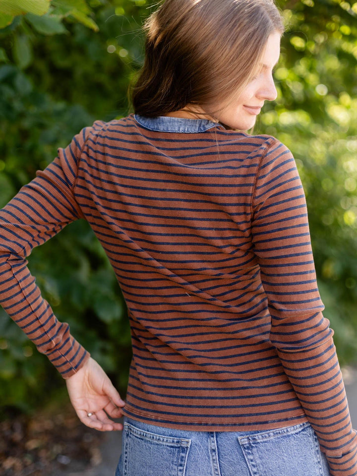A woman wearing a rust-brown and navy blue horizontally striped long-sleeve shirt with a denim collar, photographed outdoors against a blurred green foliage background.