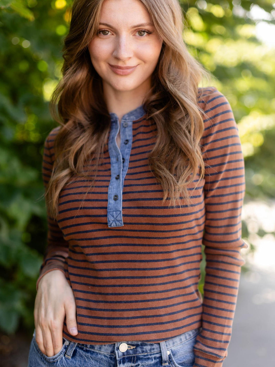 A woman wearing a rust-brown and navy blue striped long-sleeve henley shirt with a blue denim-style placket, paired with blue jeans, standing outdoors against a blurred green foliage background.