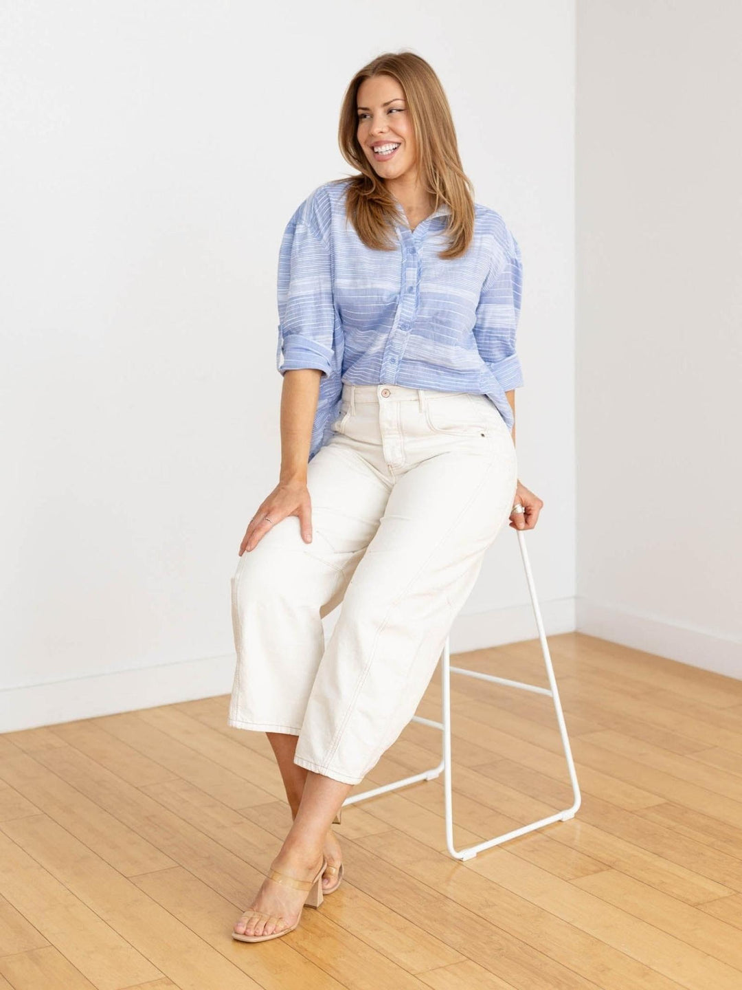 Woman in a blue striped shirt and white pants standing in a minimalistic room with a white wall and wooden floor.