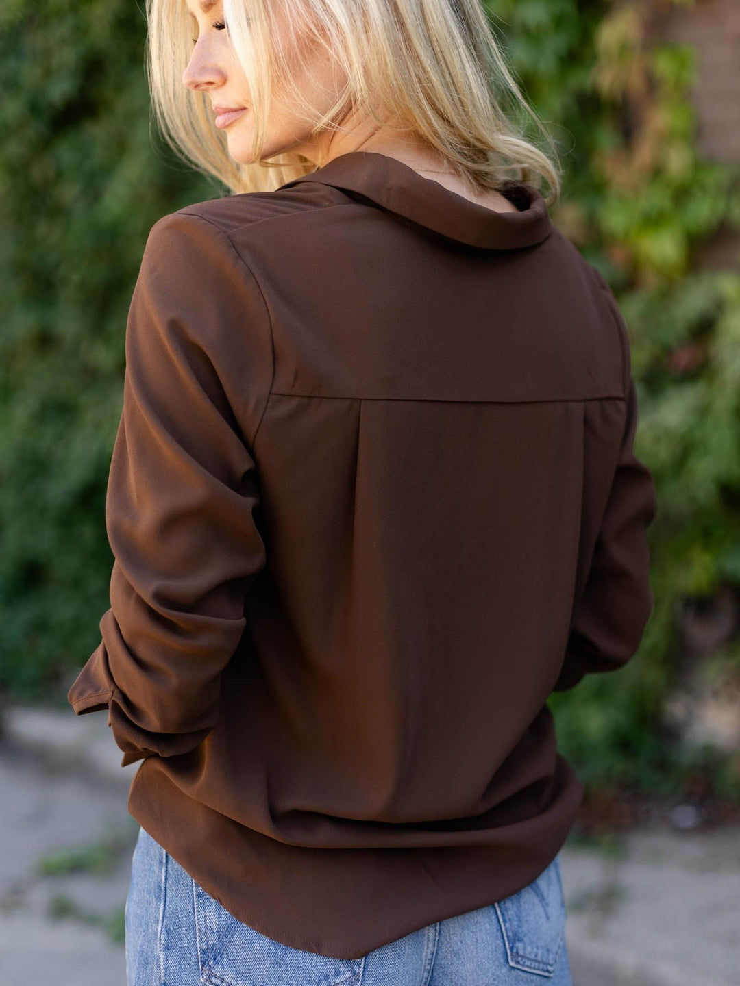 A woman wearing a brown long-sleeve collared blouse, shown from the back, paired with light blue jeans against a blurred outdoor background of green foliage.