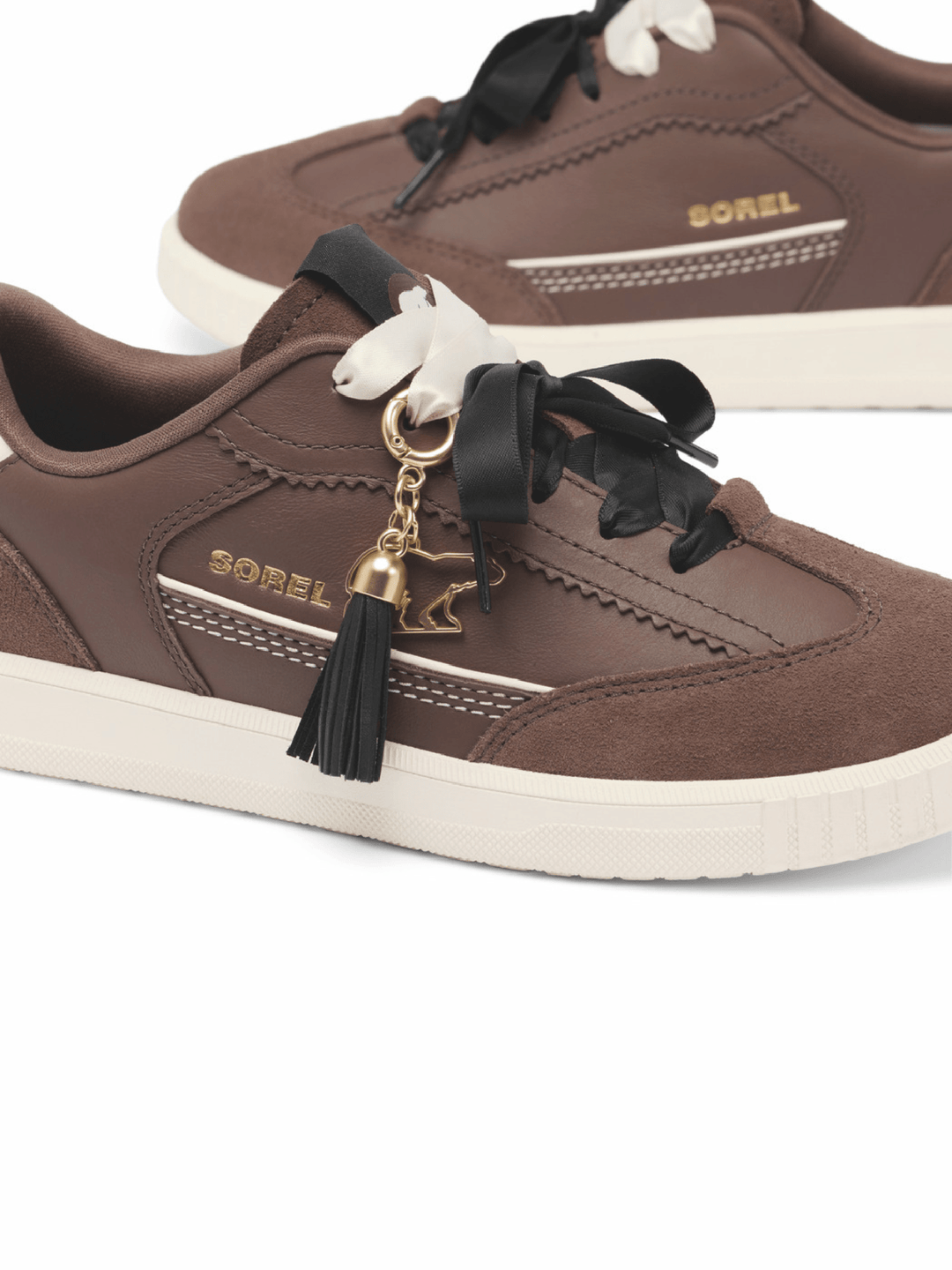 A pair of Sorel brand low-top sneakers in brown suede and leather, featuring black ribbon laces, a white horse-shaped charm, a black tassel, and gold branding.