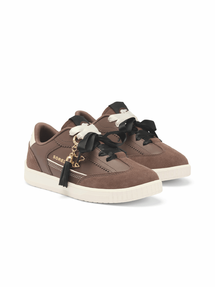 A pair of brown suede sneakers with black and white decorative laces, featuring a gold-tone tassel charm and &