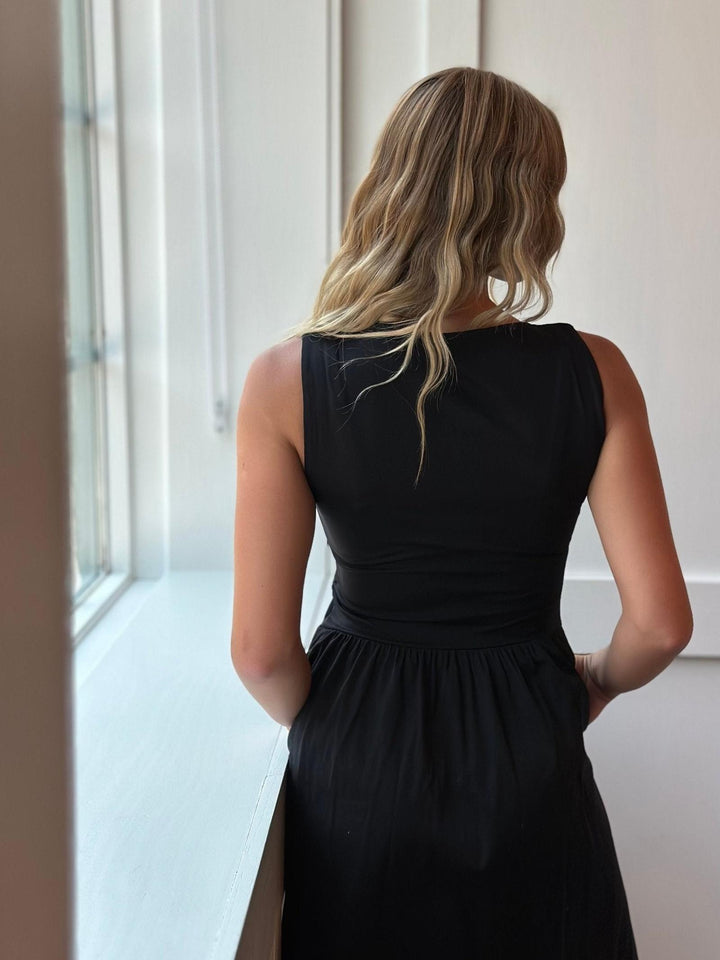 A woman with blonde wavy hair is shown from behind wearing a sleeveless black dress, standing near a bright window in a minimalist indoor setting.