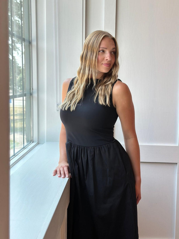 A woman with blonde hair wearing a sleeveless black midi dress, standing next to a window in a bright room.