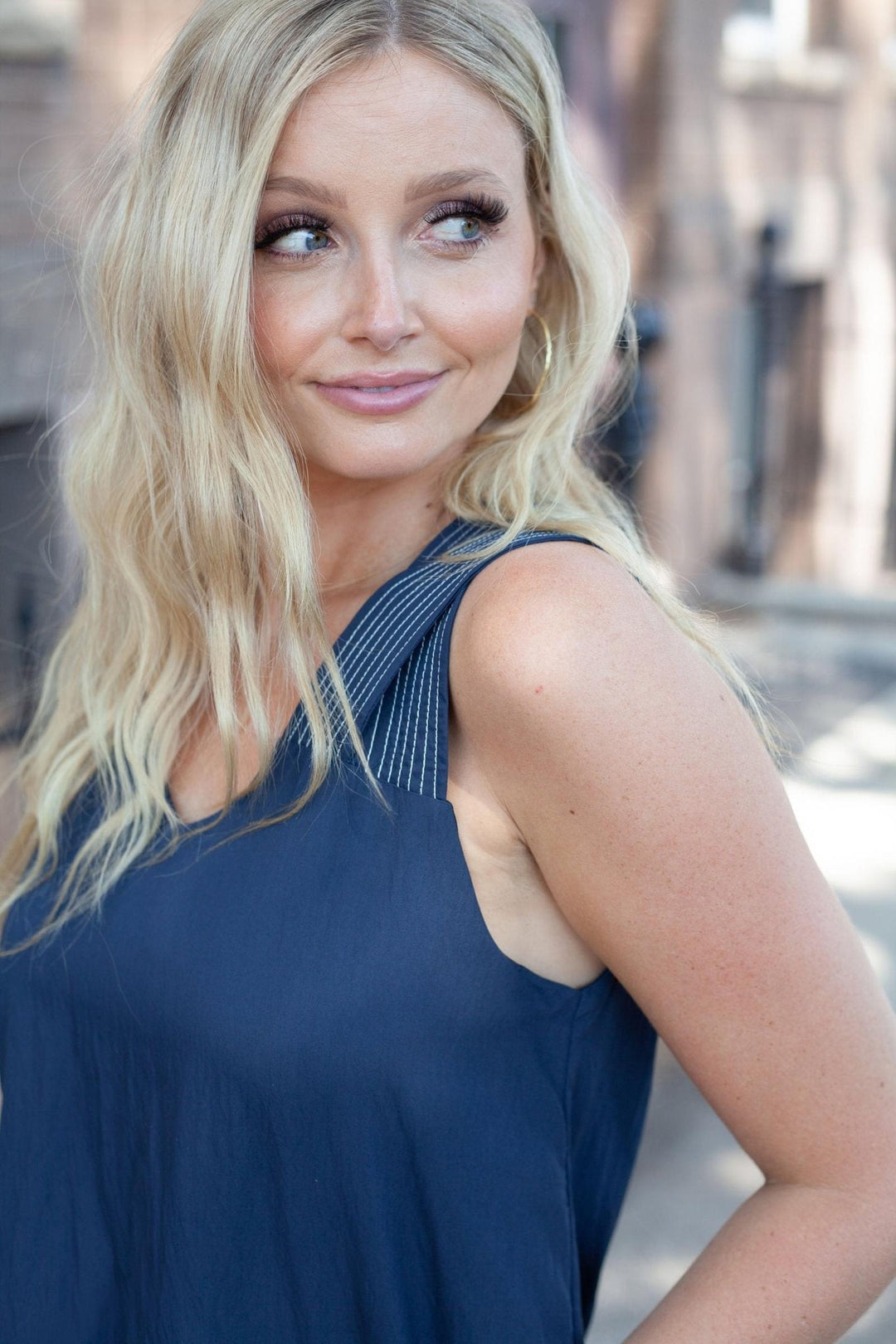 A blonde woman wearing a navy blue sleeveless top, photographed outdoors with a blurred urban background.