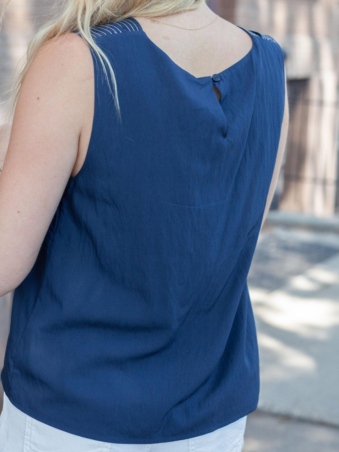 A woman wearing a sleeveless navy blue top, shown from the back. The top has a V-neckline with a small button detail at the center back and subtle decorative stitching near the shoulder straps.