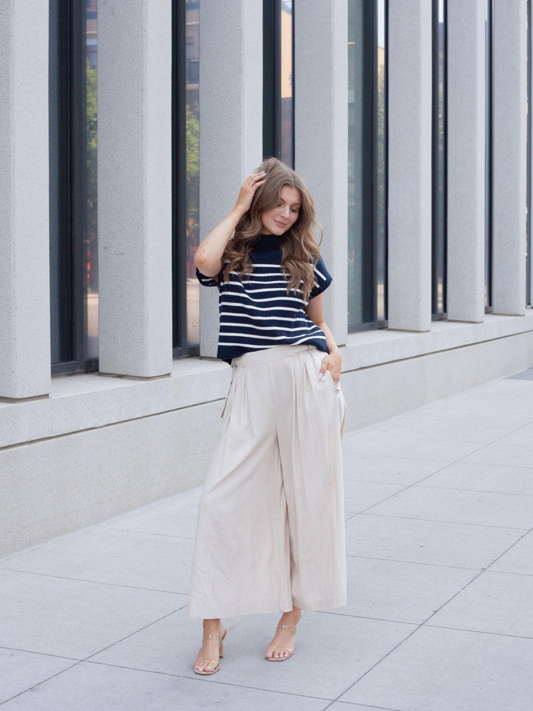 A woman stands outdoors in front of a modern building with large gray columns and windows, wearing a navy and white striped short-sleeve top with cream wide-leg cropped pants and nude heeled sandals.