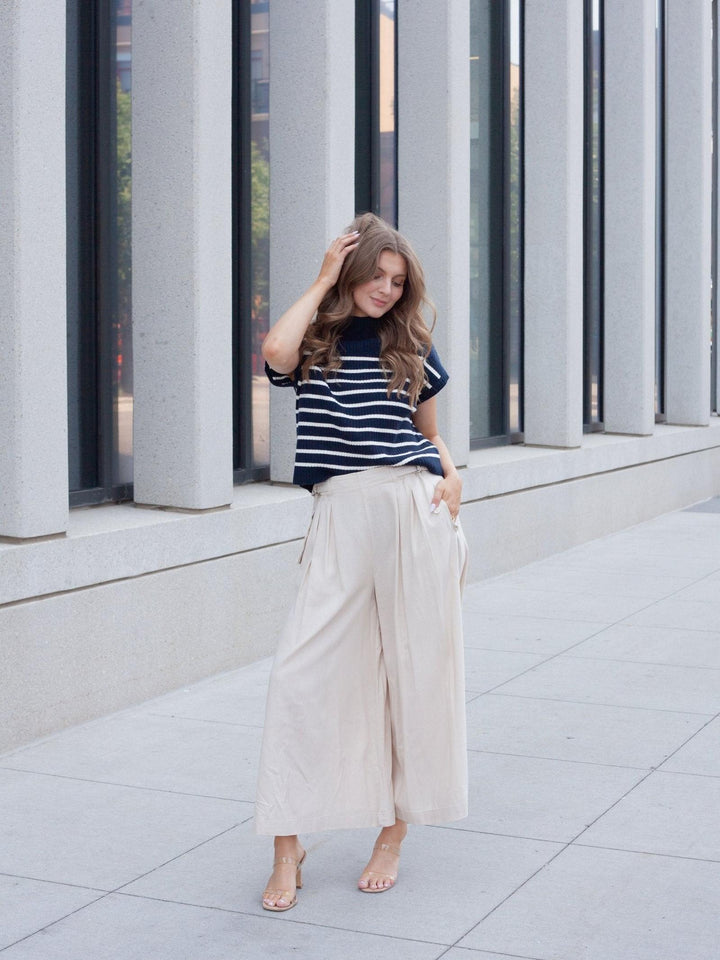 A woman stands outdoors in front of a modern building with large gray columns and windows, wearing a navy and white striped short-sleeve top with cream wide-leg cropped pants and nude heeled sandals.