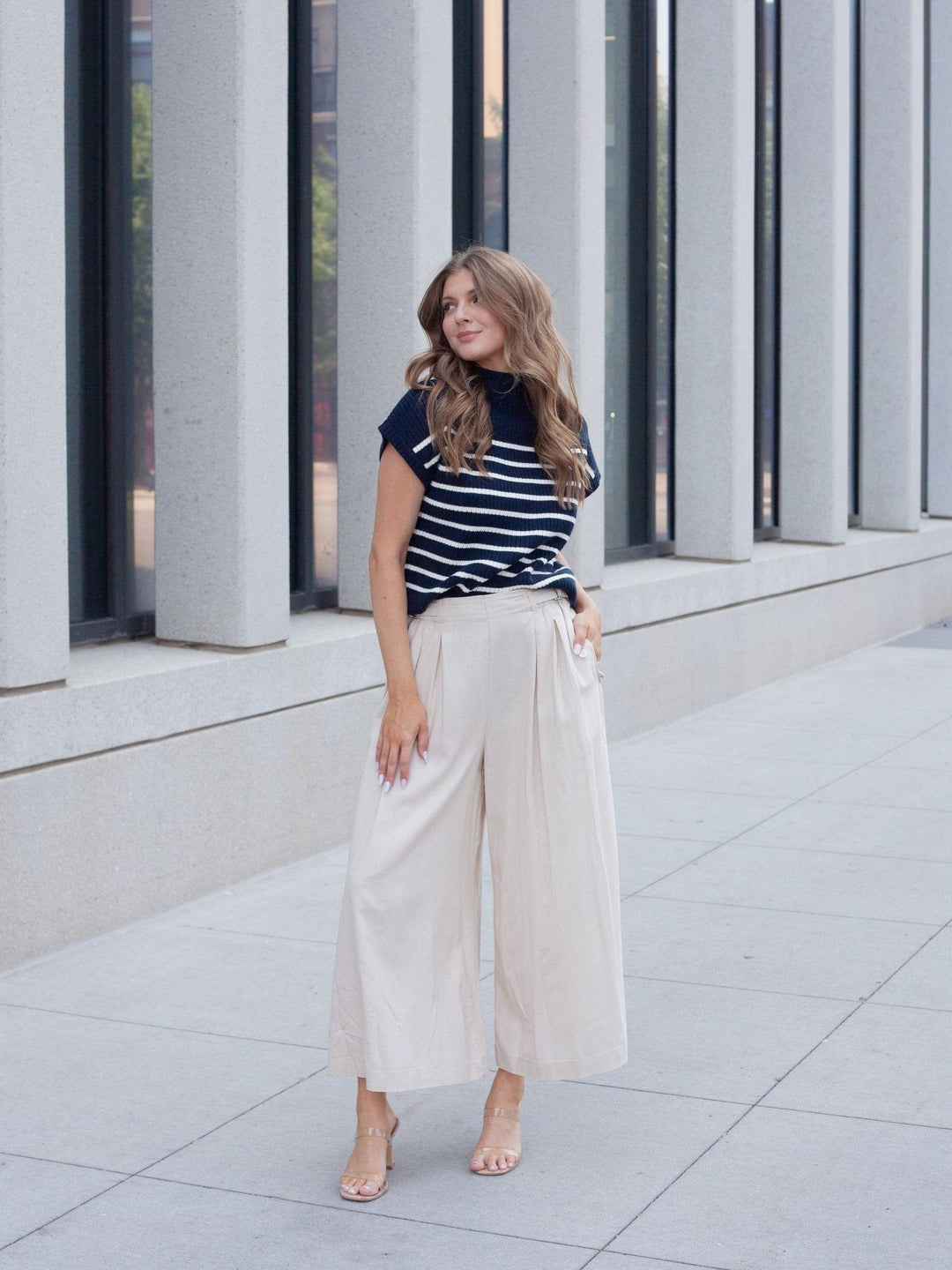 A woman stands outdoors in front of a modern building with large gray columns, wearing a navy and white horizontally striped short-sleeve top with cream wide-leg cropped pants and nude heels.