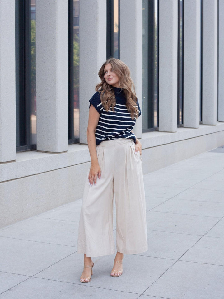 A woman stands outdoors in front of a modern building with large gray columns, wearing a navy and white horizontally striped short-sleeve top with cream wide-leg cropped pants and nude heels.
