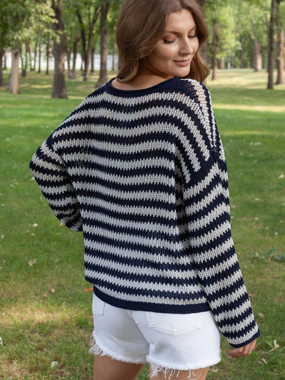 A woman wearing a navy blue and off-white horizontally striped knit sweater, shown from the back in an outdoor park setting with green grass and trees.
