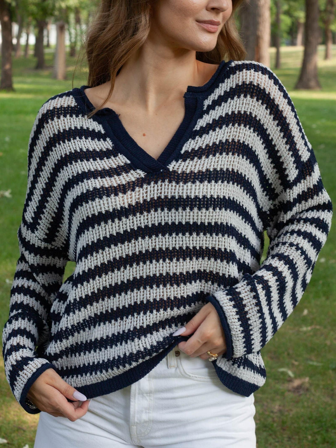 A woman wearing a navy blue and off-white striped knit sweater with a split V-neck, standing outdoors in a park with trees and grass in the background.