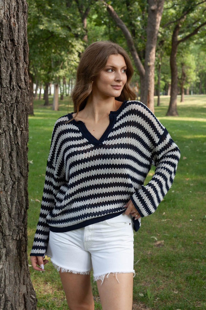 A woman wearing a black and white striped knit sweater and white denim shorts stands outdoors in a park next to a tree trunk.