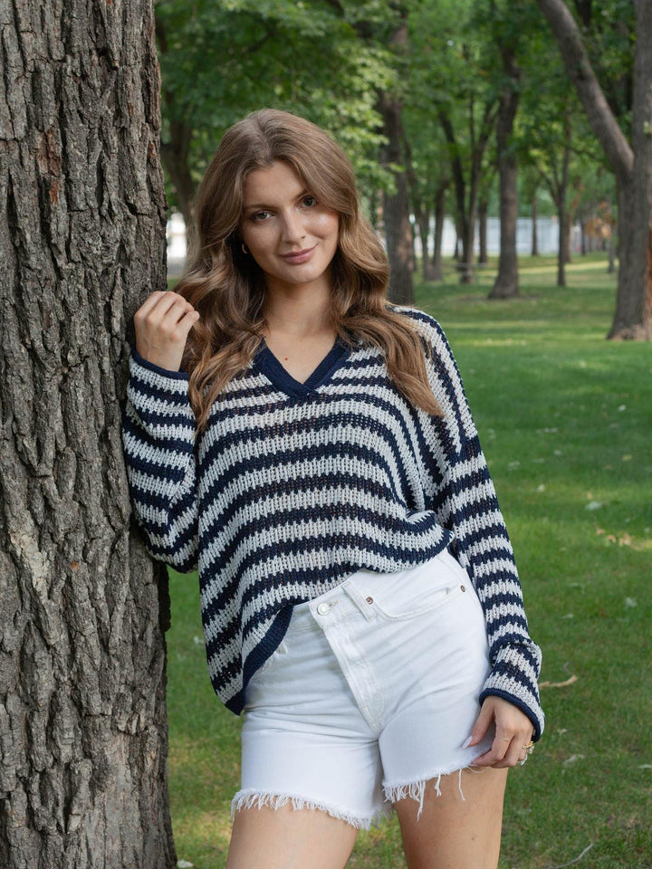 A woman wearing a navy and white striped knit sweater and white denim shorts leans against a tree in a park setting.